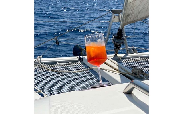 Wijnglas met oranje-rode drank op het dek van een zeilboot, net, touwen, tuigage en open zee op de achtergrond