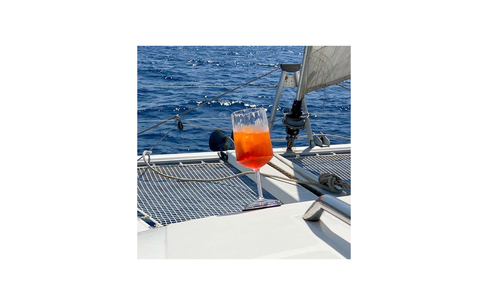 Wijnglas met oranje-rode drank op het dek van een zeilboot, net, touwen, tuigage en open zee op de achtergrond