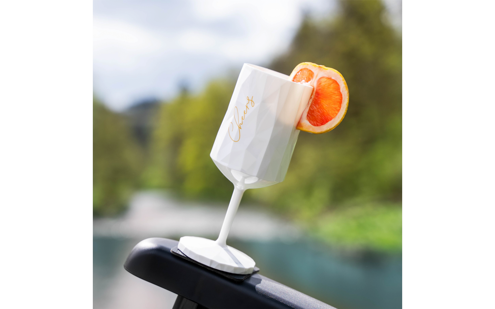 Weißer, facettierter Stielbecher mit goldener Schrift "Cheers" und halber rosa-oranger Zitrusscheibe am Rand, auf einer schwarzen Fläche vor einem verschwommenen Fluss und Bäumen