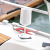 Weißer, facettierter Becher auf Stiel mit quadratischer Basis auf dem Deck eines Bootes; Reling, rote Taue und Wasser im Hintergrund