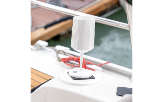 Weißer, facettierter Becher auf Stiel mit quadratischer Basis auf dem Deck eines Bootes; Reling, rote Taue und Wasser im Hintergrund