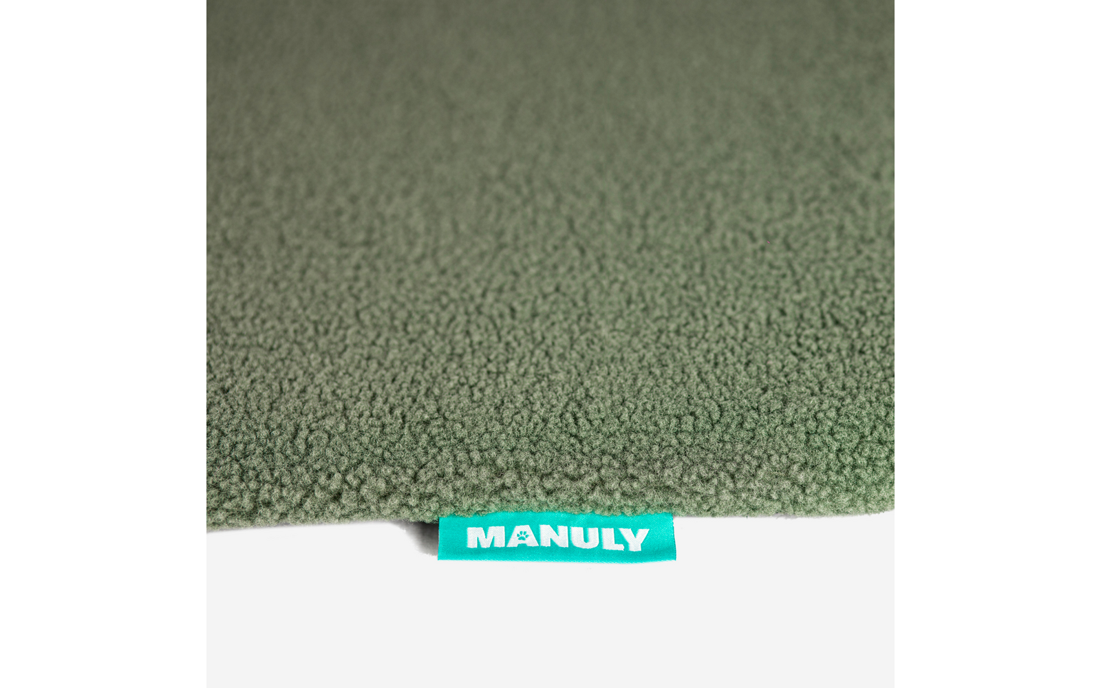 Nahaufnahme eines strukturierten olivgrünen Textils mit einer türkisen Stofflasche mit weißer Aufschrift MANULY am unteren Rand und einem hellen Hintergrund darunter