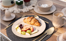Assiette de petit-déjeuner avec croissant, fruits et fromage, à côté d'une tasse de café, coquille d'œuf, bol et tasses supplémentaires sur une table dressée