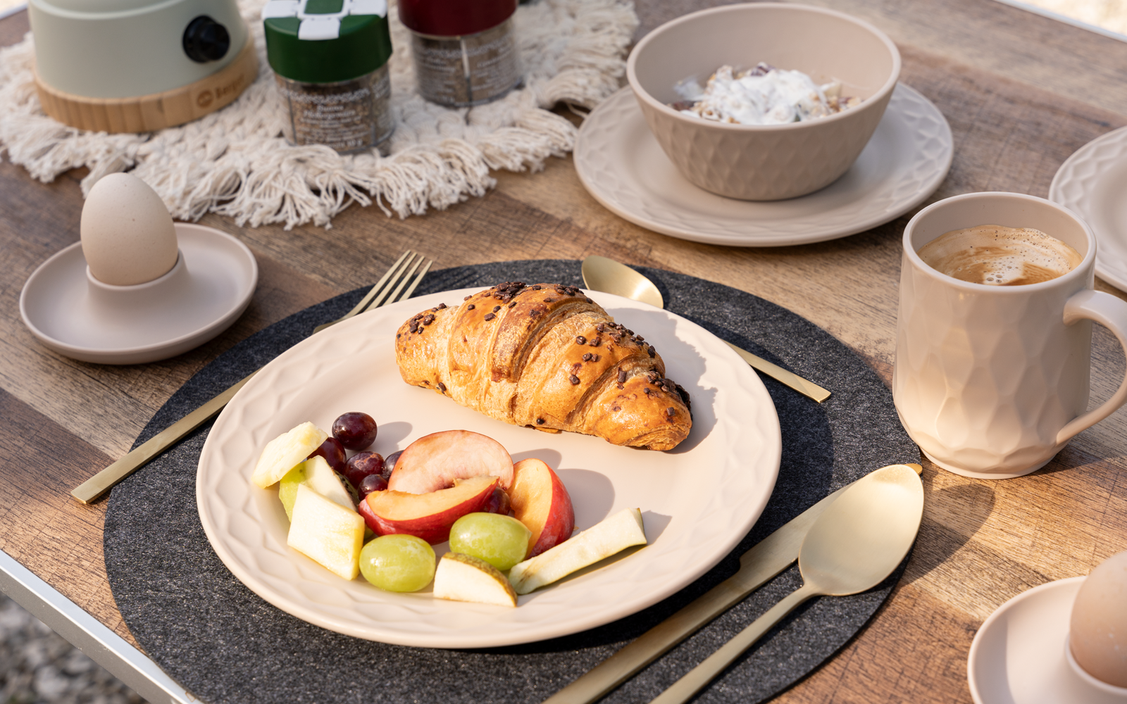 Assiette de petit-déjeuner avec croissant, fruits et fromage, à côté d'une tasse de café, coquille d'œuf, bol et tasses supplémentaires sur une table dressée