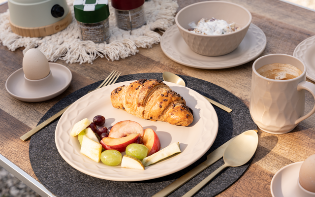 Assiette de petit-déjeuner avec croissant, fruits et fromage, à côté d'une tasse de café, coquille d'œuf, bol et tasses supplémentaires sur une table dressée