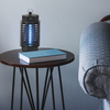 Blauer Insektenvernichter auf einem dunklen runden Tisch mit Buch, daneben ein grauer Stuhl und ein Ventilator