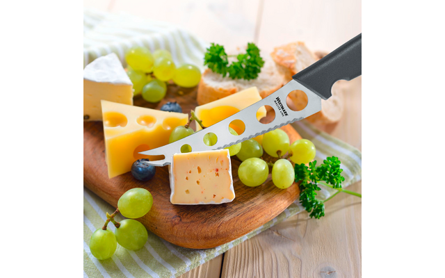 Holzbrett mit mehreren Käsestücken, grünen Trauben, einer Blaubeere, Petersilienzweigen, Brotscheiben und einem Käsemesser mit Löchern