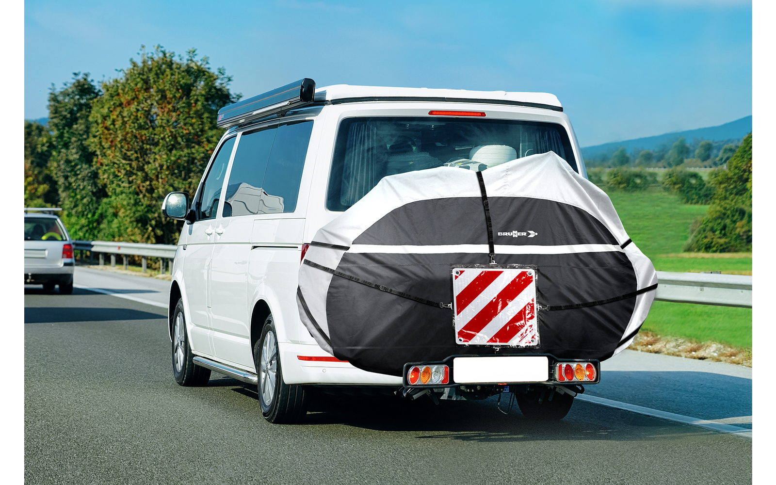 Furgone bianco che percorre una strada di campagna. Sul retro una copertura rotonda nera con un pannello di avvertimento rosso e bianco diagonale fissato. Fanali posteriori e portapacchi sul tetto visibili. Campi verdi e alberi ai bordi della strada, cielo azzurro