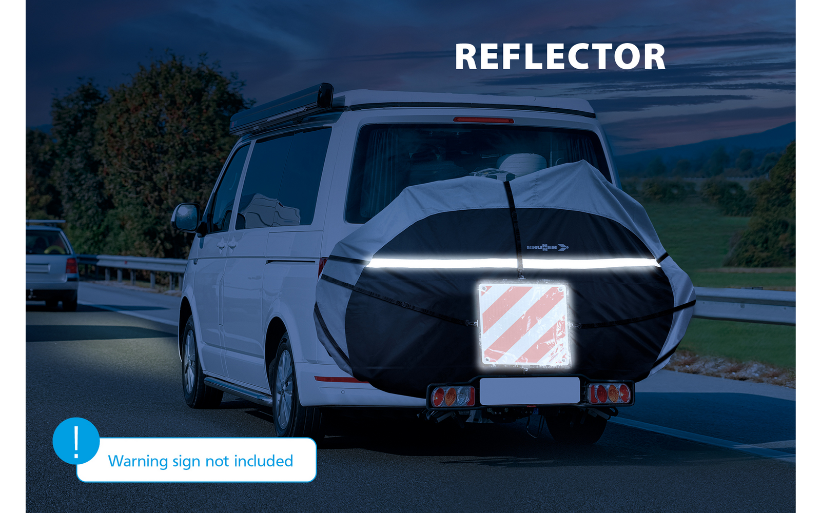 Furgone bianco su strada di campagna al crepuscolo, vista posteriore con ruota di scorta in copertura nera con fascia riflettente orizzontale e targa a strisce diagonali rosse e bianche; altra auto visibile dietro a sinistra, guardrail, alberi e colline. Testo visibile: "REFLECTOR" e "Warning sign not included"