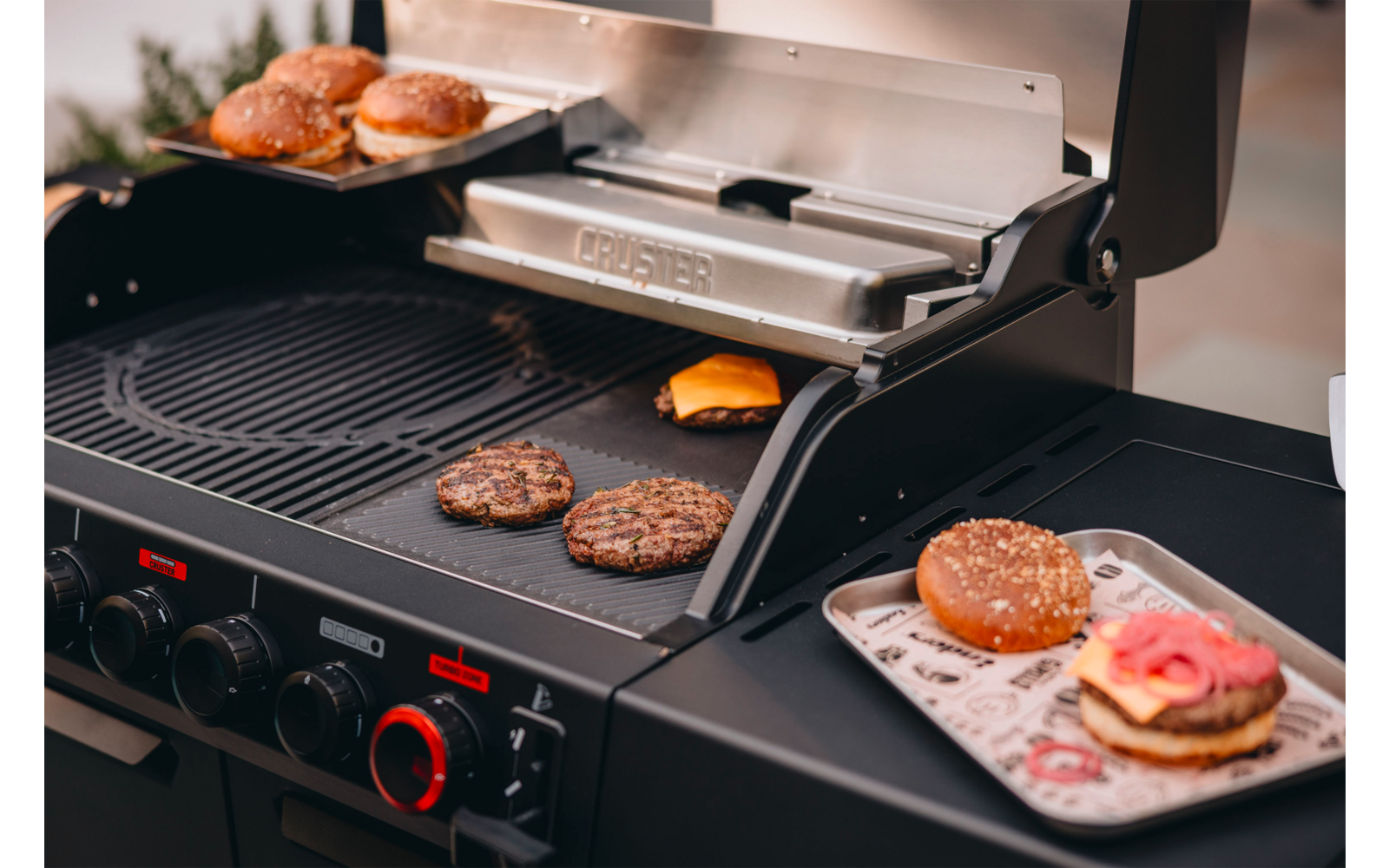 Barbecue à gaz avec couvercle ouvert, deux galettes de hamburger et une tranche de fromage sur la grille, moitiés de petits pains sur une étagère et un hamburger prêt sur une assiette