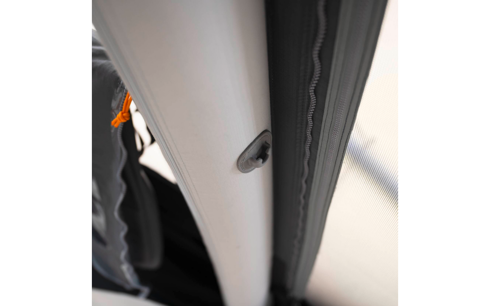 Nahaufnahme zweier vertikaler Rohre (weiß und grau) mit grauem Plastikclip am weißen Rohr, rechts Reißverschluss und Stoffkante, links schwarzer Stoff und orangefarbene Schnur