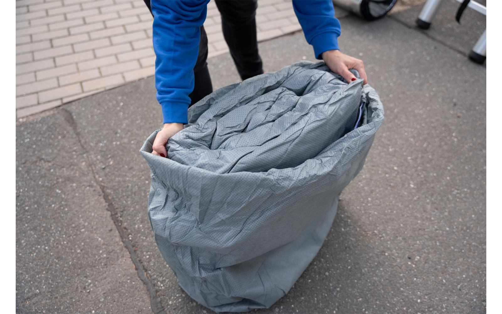 Persoon in een blauw sweatshirt en zwarte broek houdt een open grijze zak met opgerold grijs materiaal; asfalt en een betegeld trottoir zichtbaar