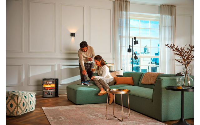 Living room with green sectional sofa, two people (one seated, one standing), small modern heater with visible flames on the left, two round side tables, area rug, window with city view, wall paneling, curtains and a vase with branches