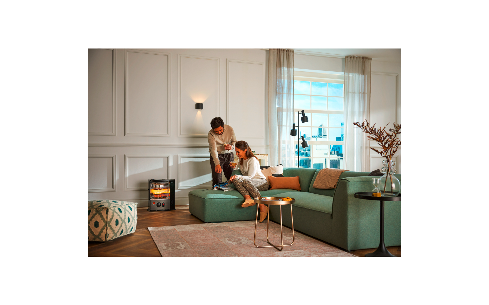 Living room with green sectional sofa, two people (one seated, one standing), small modern heater with visible flames on the left, two round side tables, area rug, window with city view, wall paneling, curtains and a vase with branches