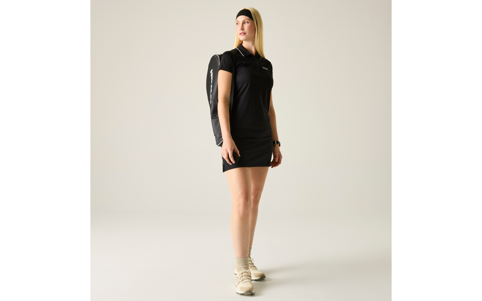 Person steht vor hellem Hintergrund, trägt schwarzes Polohemd, kurzen schwarzen Rock, schwarzen Rucksack, schwarzes Stirnband, beige Socken und beige Sneaker, blickt zur Seite