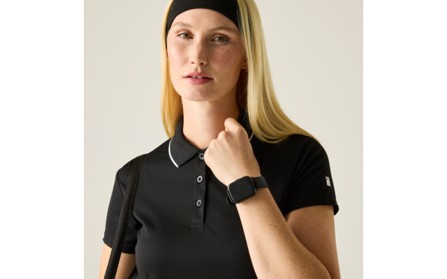 Person mit langen blonden Haaren und schwarzem Stirnband, trägt schwarzes Polohemd mit weißer Kantennaht, schwarze Smartwatch am Handgelenk, hält den Kragen; heller einfarbiger Hintergrund