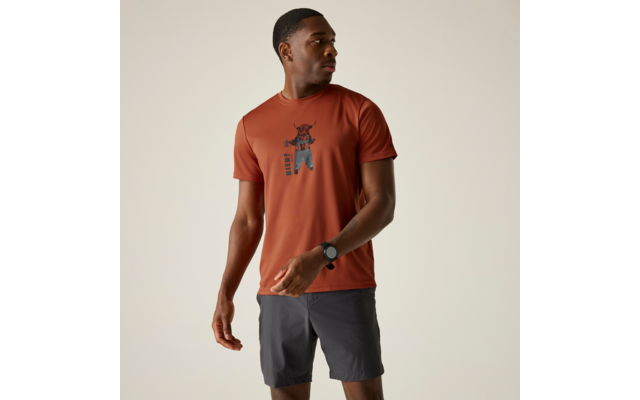 Person steht vor hellem Hintergrund und blickt nach links, trägt rostfarbenes T‑Shirt mit schwarzem zentralem Aufdruck, graue Shorts und eine schwarze Armbanduhr am linken Handgelenk