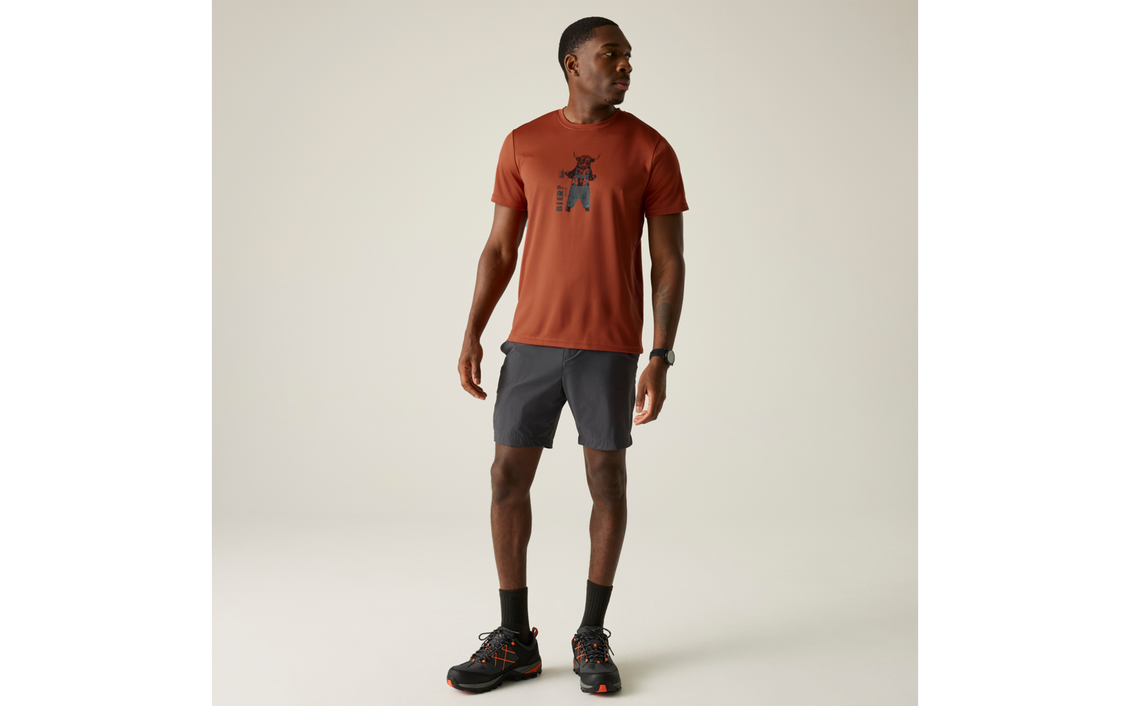 Person steht, trägt rostfarbenes T‑Shirt mit Aufdruck, dunkelgraue Shorts, schwarze Socken und schwarze Sportschuhe, blickt nach links