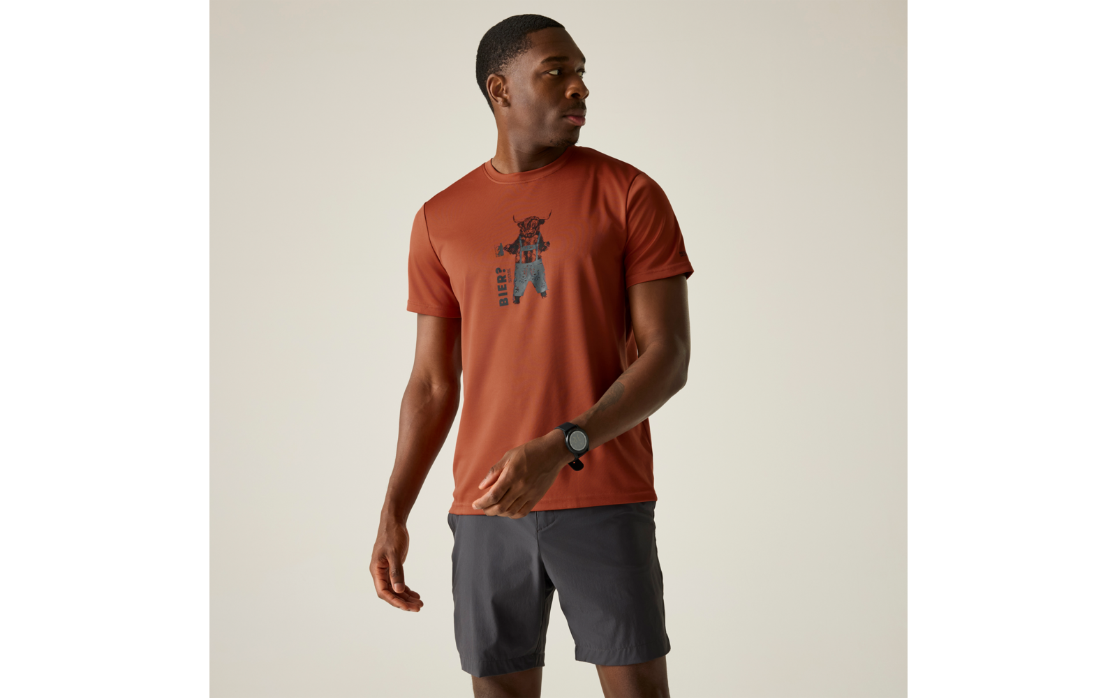 Person steht vor hellem Hintergrund und blickt nach links, trägt rostfarbenes T‑Shirt mit schwarzem zentralem Aufdruck, graue Shorts und eine schwarze Armbanduhr am linken Handgelenk