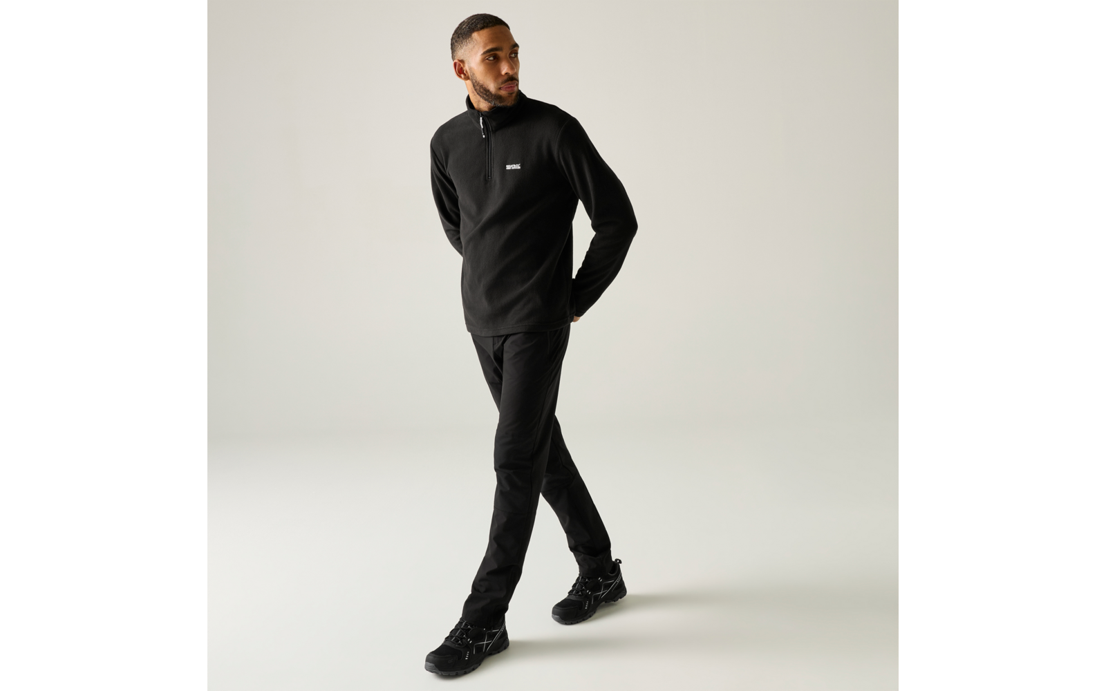 Man in zwarte trui en zwarte broek met zwarte sneakers, loopt met gekruiste benen voor een lichte achtergrond, handen achter de rug, kijkt naar rechts