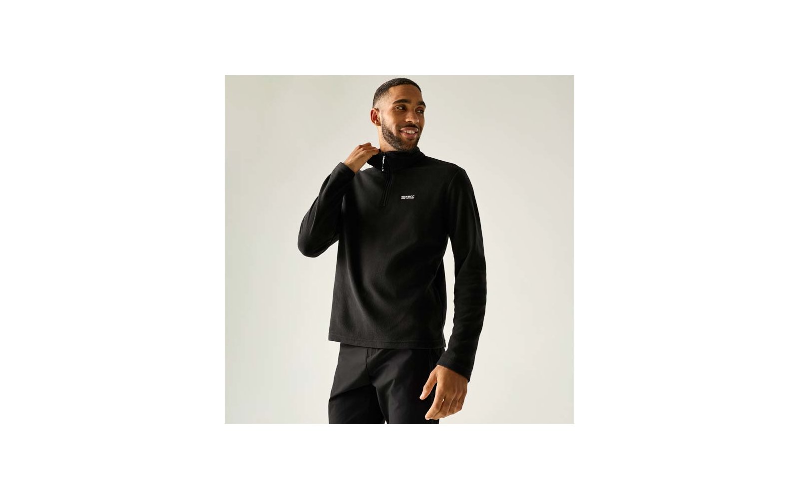 Man staand met een zwarte trui met korte rits bij de kraag en een klein logo op de borst, rechterhand bij de kraag