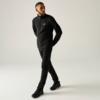 Man in zwarte trui en zwarte broek met zwarte sneakers, loopt met gekruiste benen voor een lichte achtergrond, handen achter de rug, kijkt naar rechts