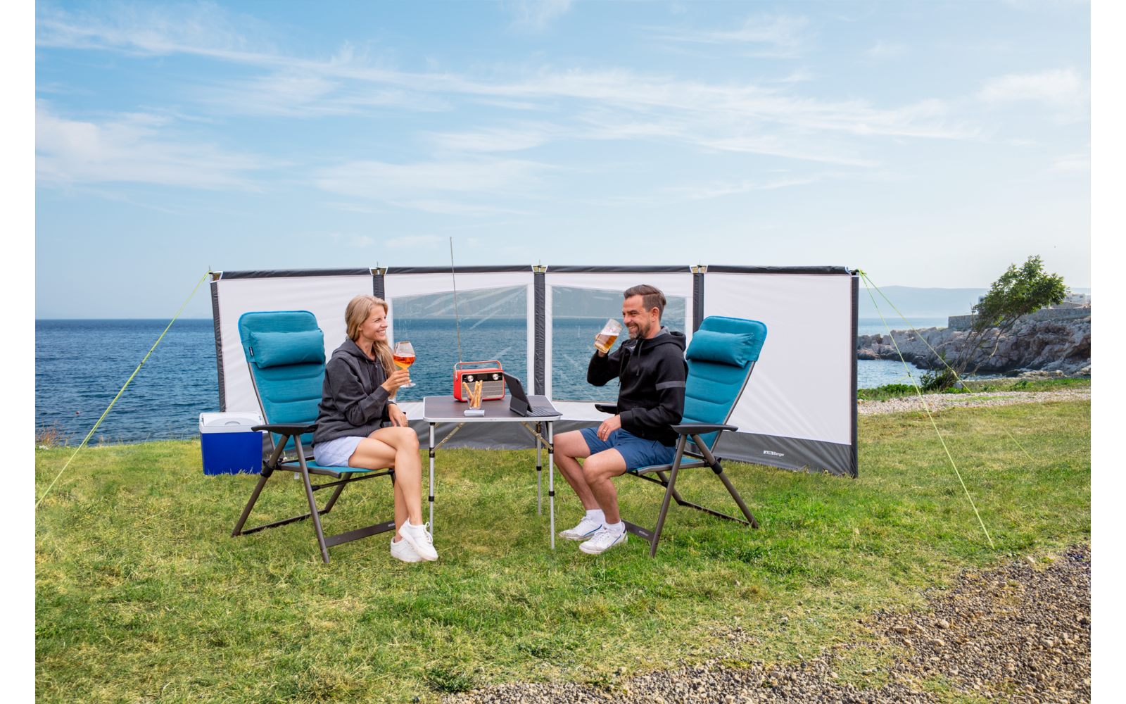 Zwei Personen sitzen an einem Campingtisch mit Faltstühlen auf einer Wiese am Meer, im Hintergrund ist eine Windschutzwand aufgestellt