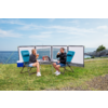 Zwei Personen sitzen an einem Campingtisch mit Faltstühlen auf einer Wiese am Meer, im Hintergrund ist eine Windschutzwand aufgestellt