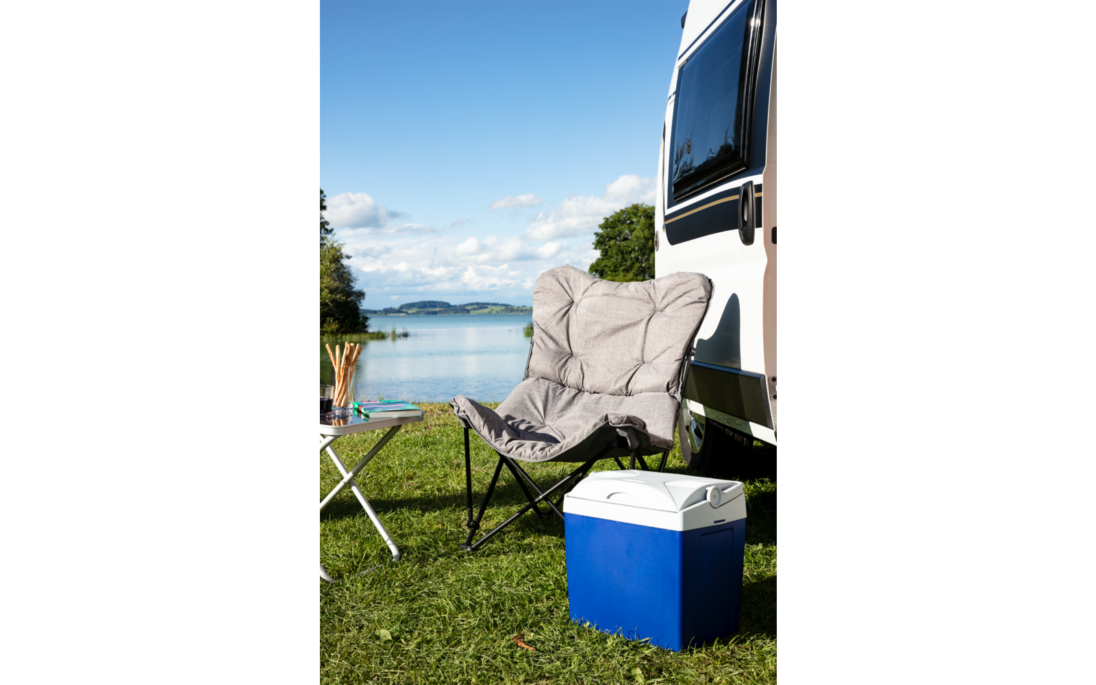 Silla plegable y nevera sobre hierba junto a una autocaravana con un lago y montañas al fondo