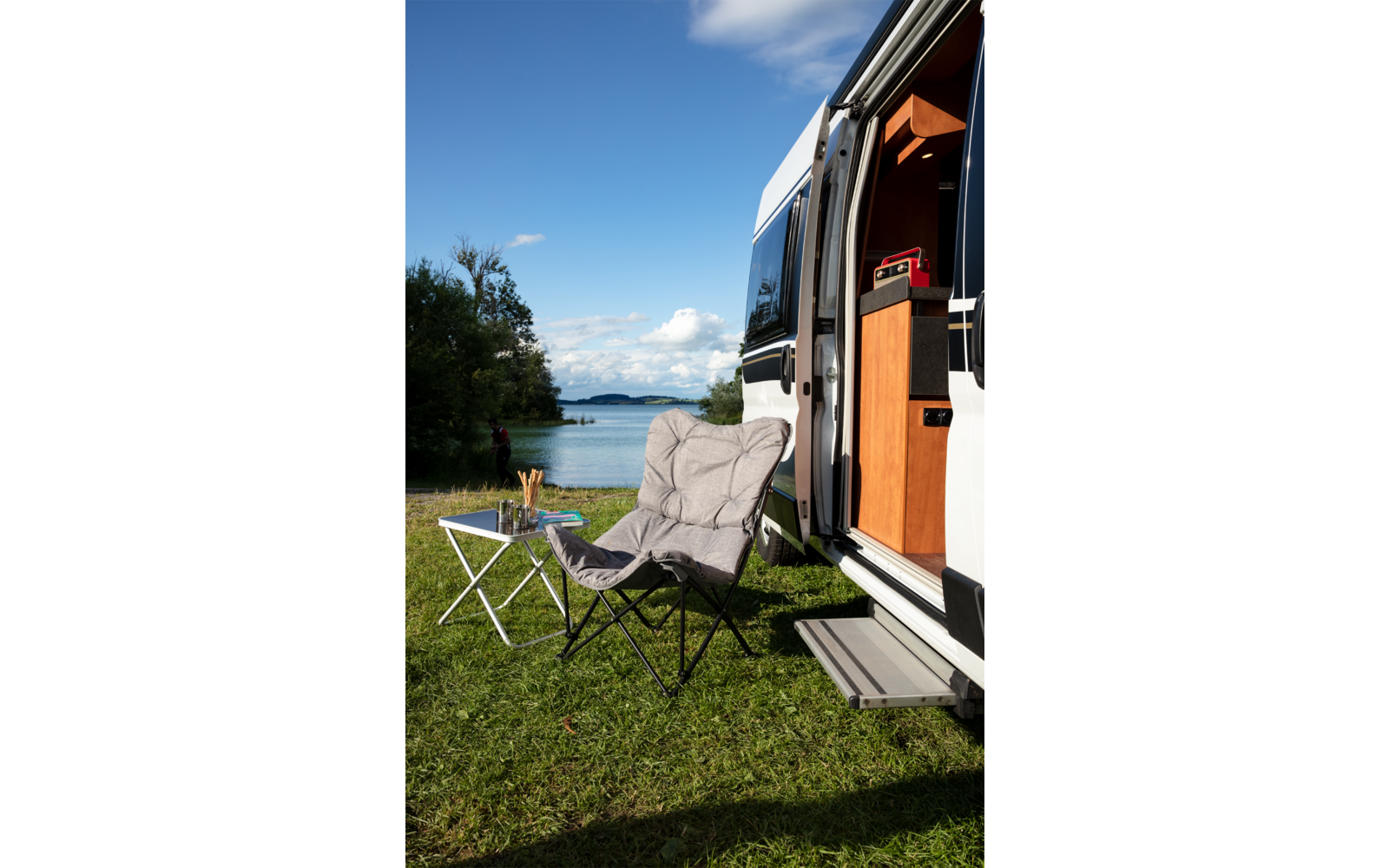 Silla de camping y pequeña mesa plegable junto a una autocaravana abierta en un campo de hierba bajo un cielo azul con nubes