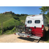Rückseite eines weißen und roten Wohnmobils mit zwei auf einem Fahrradträger montierten Fahrrädern, im Hintergrund grüne Hügel und ein blau bewölkter Himmel