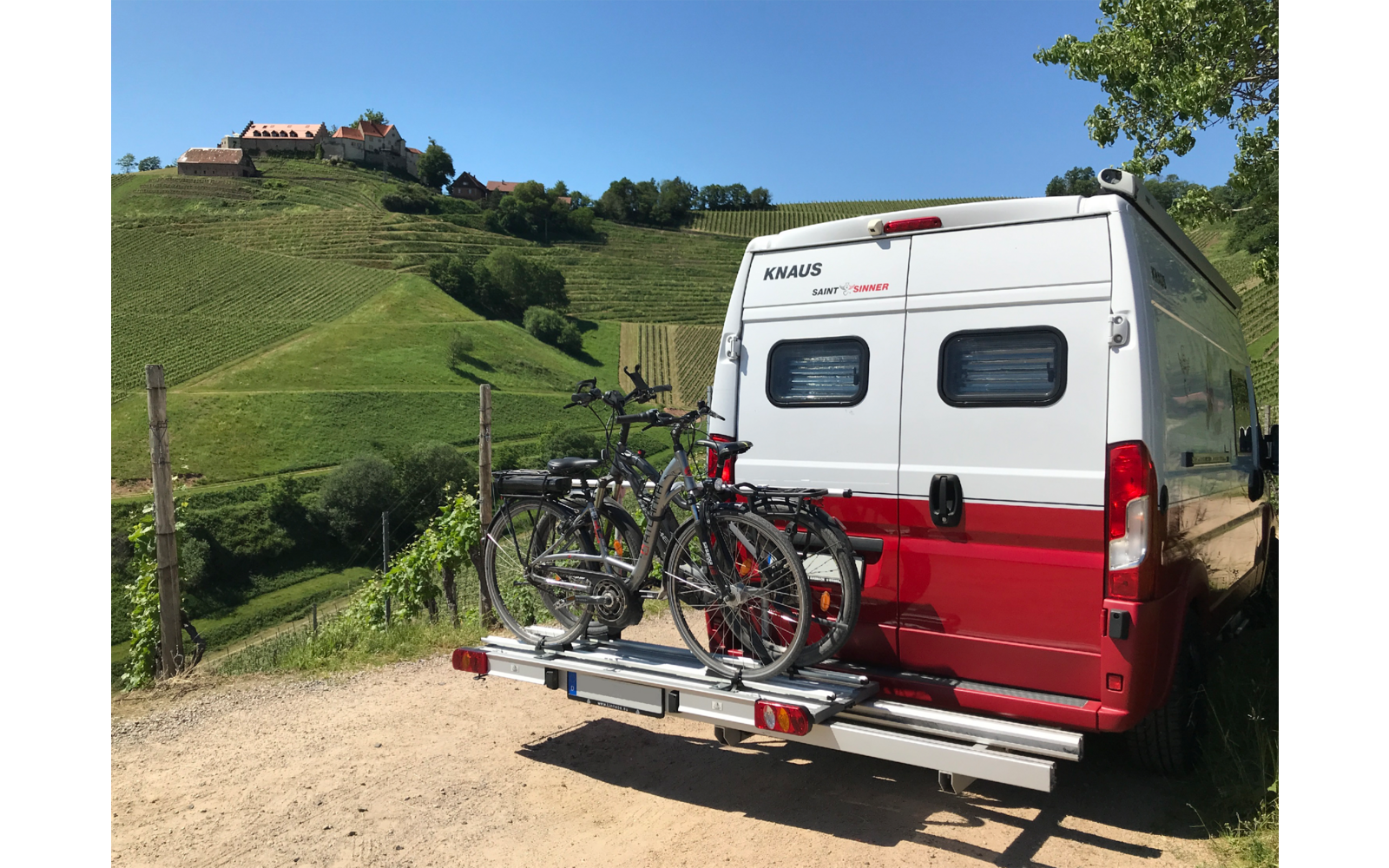 Rückseite eines weißen und roten Wohnmobils mit zwei auf einem Fahrradträger montierten Fahrrädern, im Hintergrund grüne Hügel und ein blau bewölkter Himmel