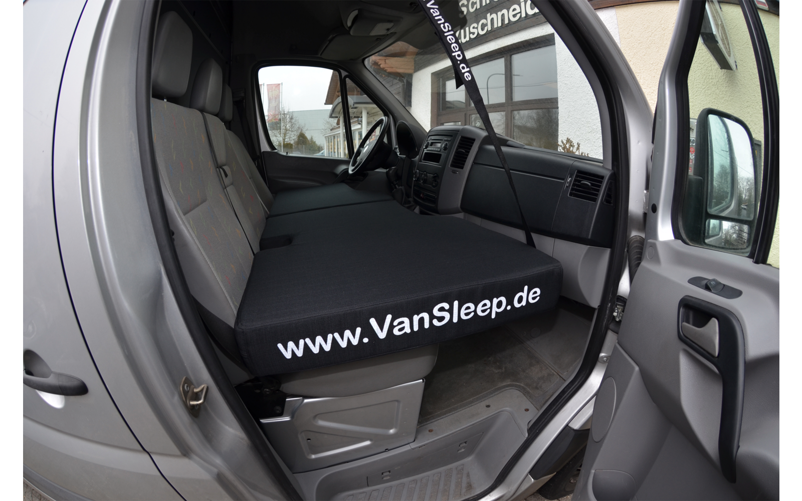 Innenansicht eines Fahrzeugs mit offener Tür, Blick auf den Fahrersitz und ein flaches Bett mit der Aufschrift www.VanSleep.de
