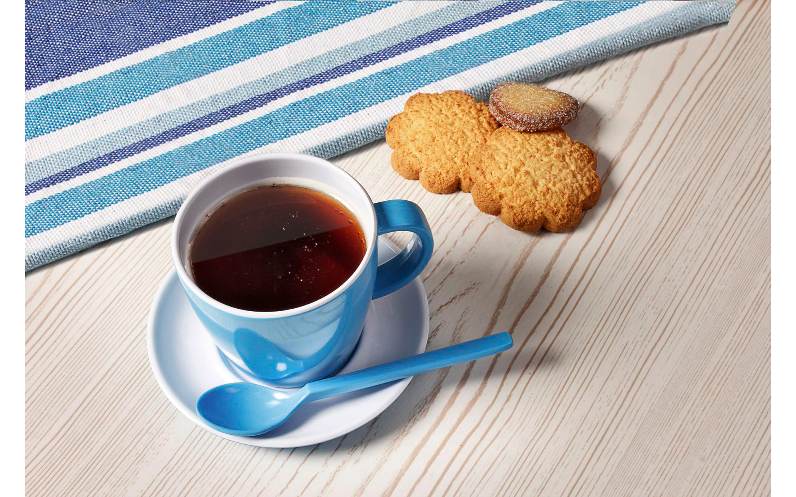 Blaue Tasse mit Tee auf einer Untertasse, daneben drei Kekse und ein Löffel auf einem hellen Tisch