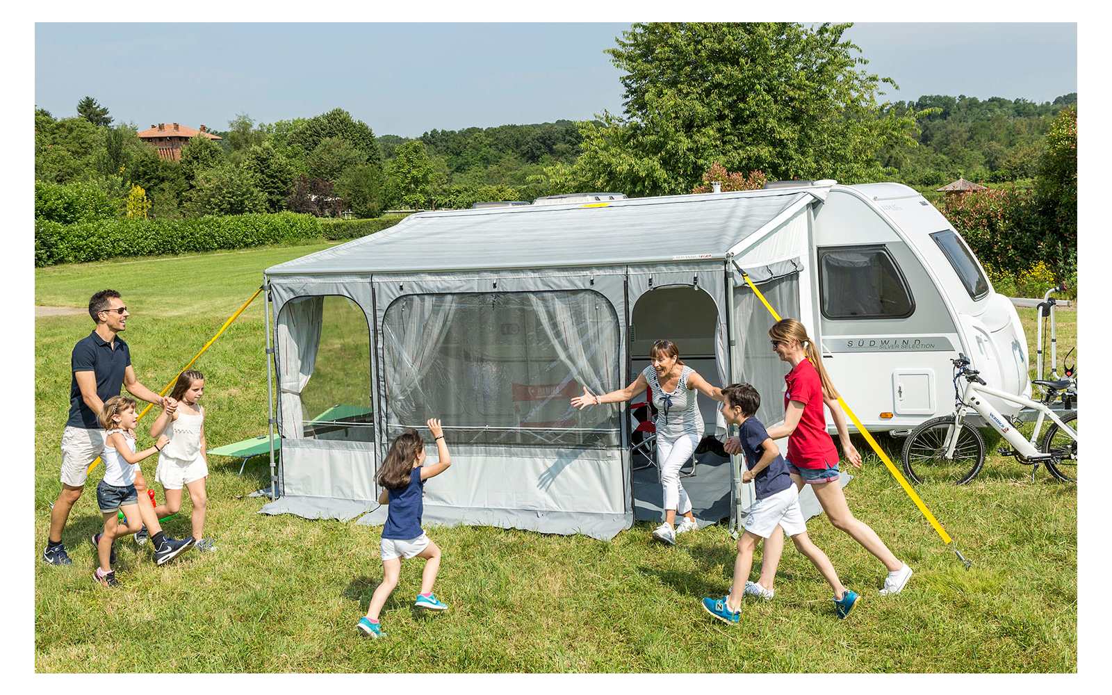 Familie mit vier Kindern spielt vor einem Wohnwagen mit angebautem Vorzelt auf einer Wiese