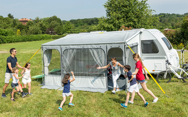 Familie mit vier Kindern spielt vor einem Wohnwagen mit angebautem Vorzelt auf einer Wiese