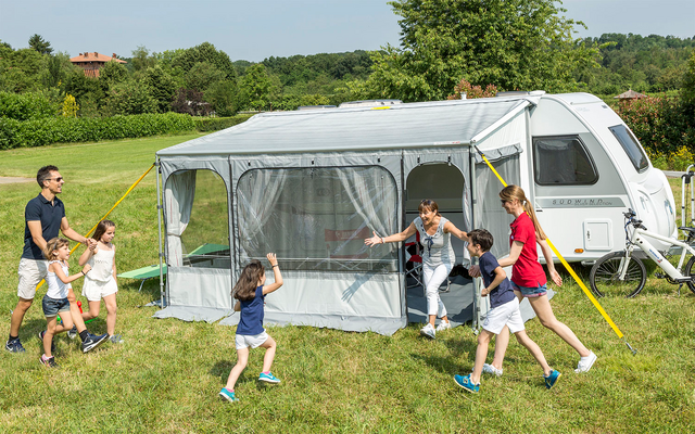 Eine Familie mit zwei Erwachsenen und drei Kindern spielt auf einer grünen Wiese vor einem Wohnwagen mit angebautem Zelt