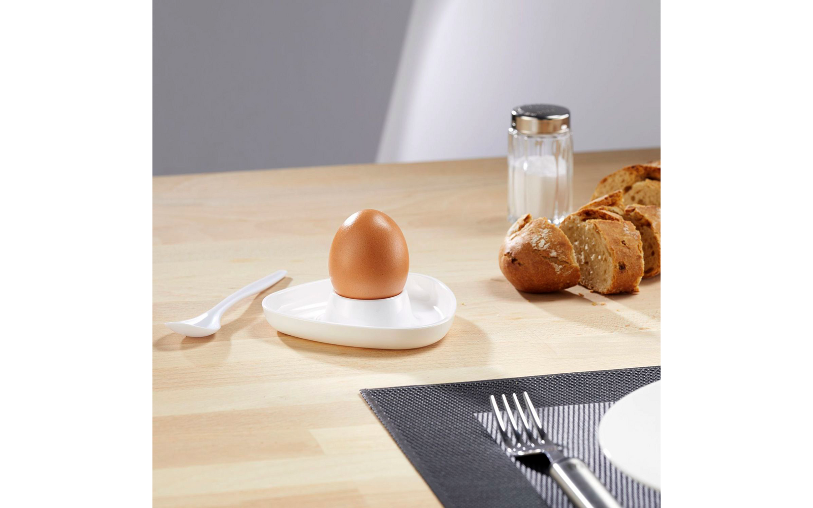 Boiled egg in white egg cup, knife, salt shaker, and bread on wooden table