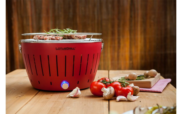 A red spherical grill on a wooden table, with fresh tomatoes, garlic cloves, and mushrooms beside it