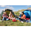 Three women sit on grass in front of two tents, eating and laughing