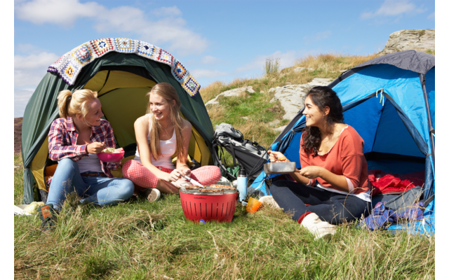 Three women sit on grass in front of two tents, eating and laughing