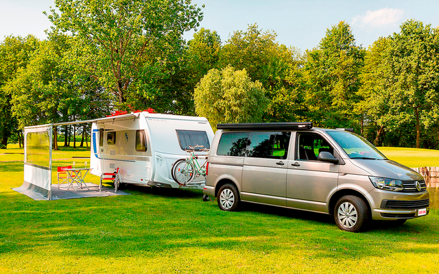 Silberner Van zieht einen weißen Wohnwagen auf einem grünen Feld mit Bäumen im Hintergrund