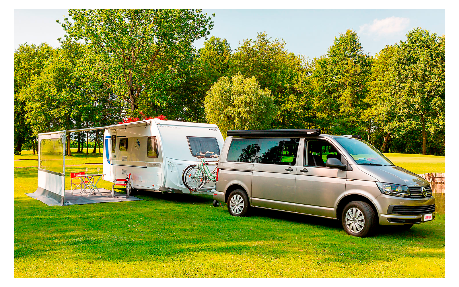Silberner Van zieht einen weißen Wohnwagen auf einem grünen Feld mit Bäumen im Hintergrund