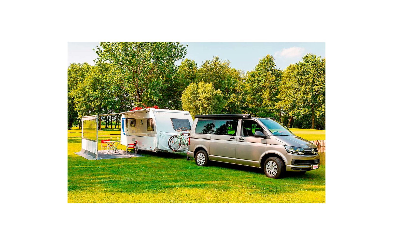 Silberner Van zieht einen weißen Wohnwagen auf einem grünen Feld mit Bäumen im Hintergrund