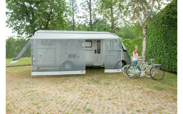 Ein geparkter Wohnwagen mit einer ausfahrbaren Markise auf einem schottrigen Untergrund, davor zwei Fahrräder, umgeben von Bäumen