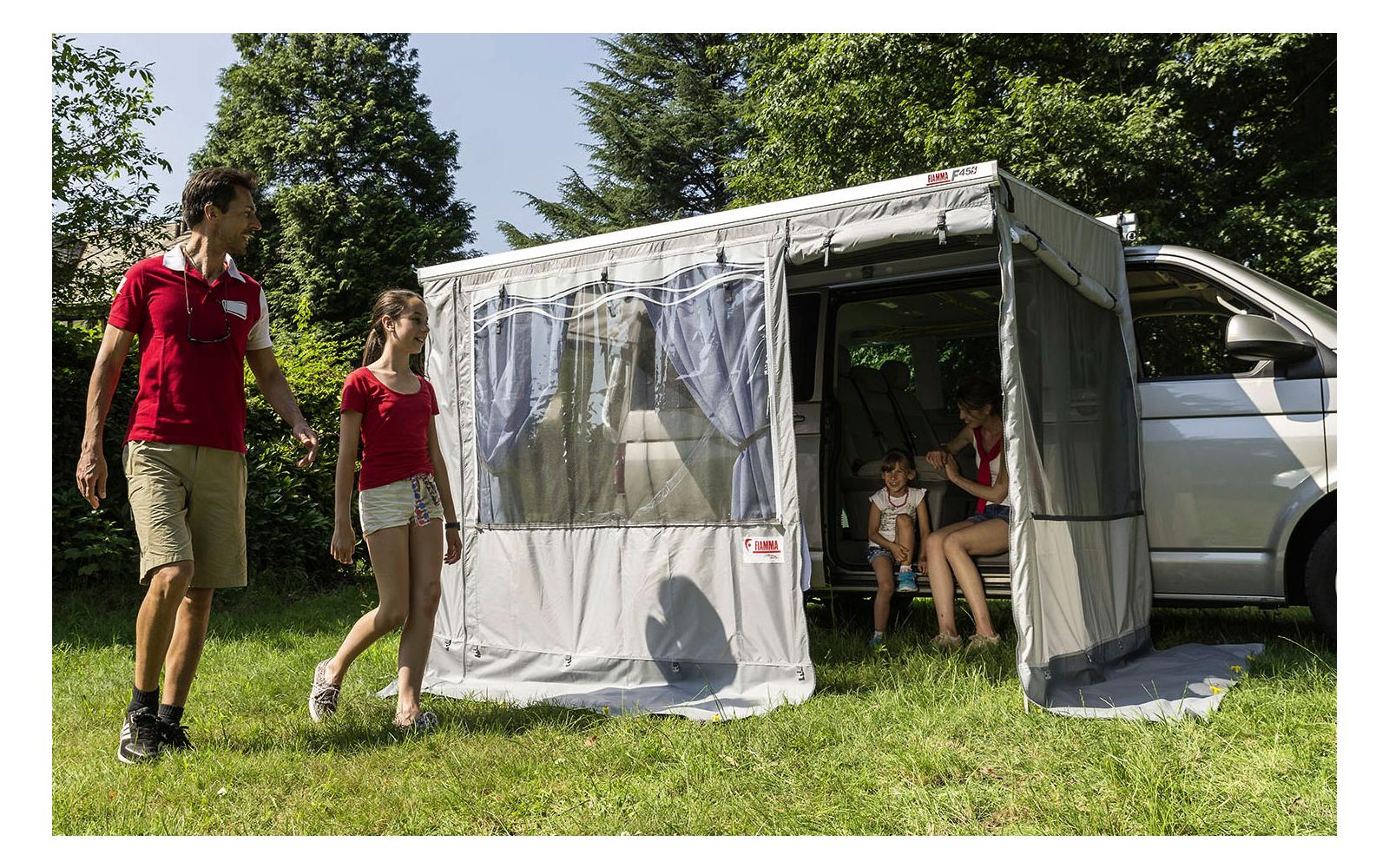 Two people walking beside a parked camper van on a grassy area with trees in the background
