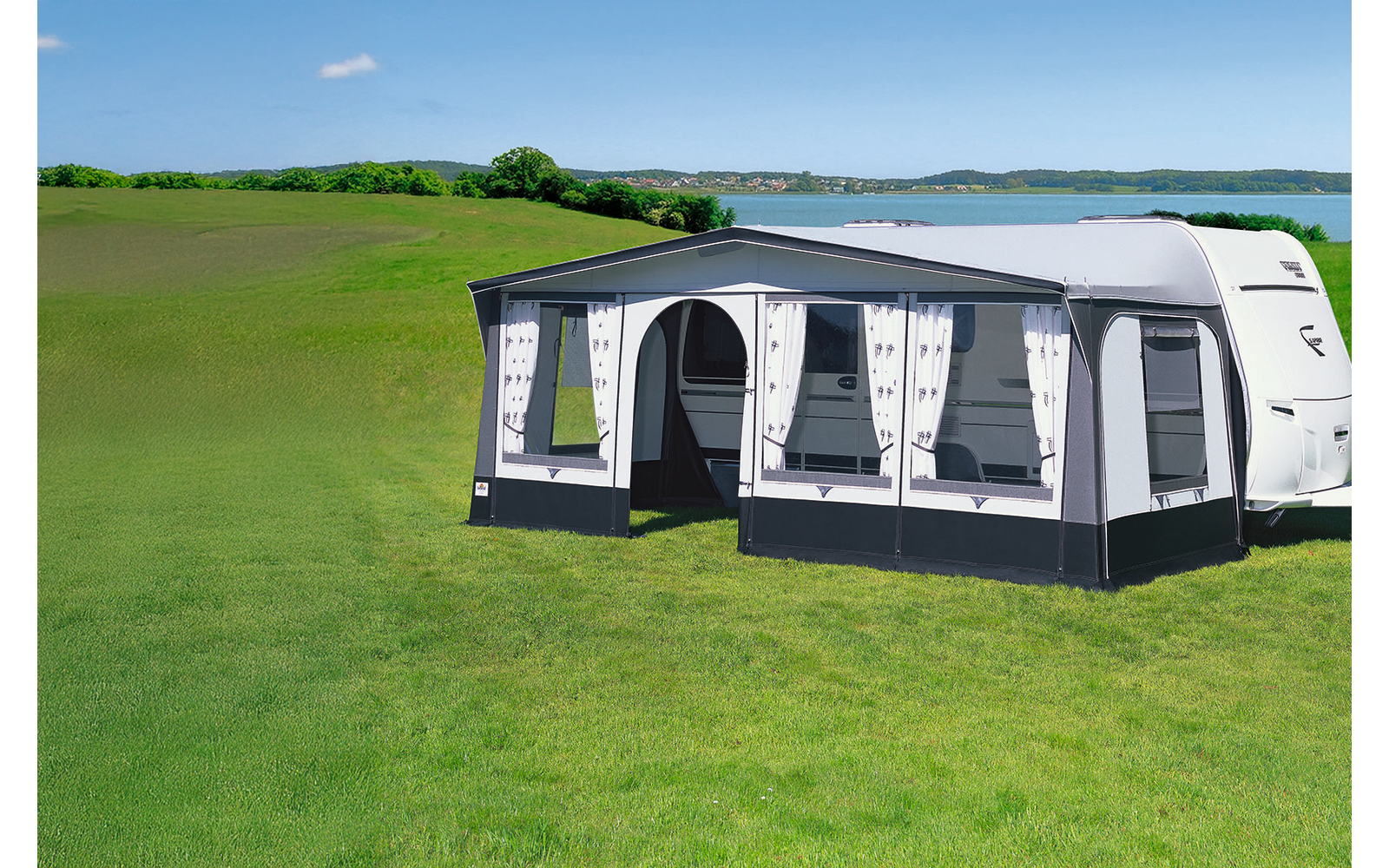 Caravan with attached bright awning on a green meadow in front of a lake and blue sky