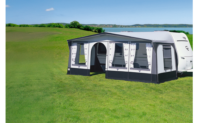 Caravan with attached bright awning on a green meadow in front of a lake and blue sky