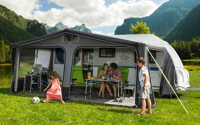 People sitting and standing in front of a caravan with an attached awning on a green meadow in the mountains