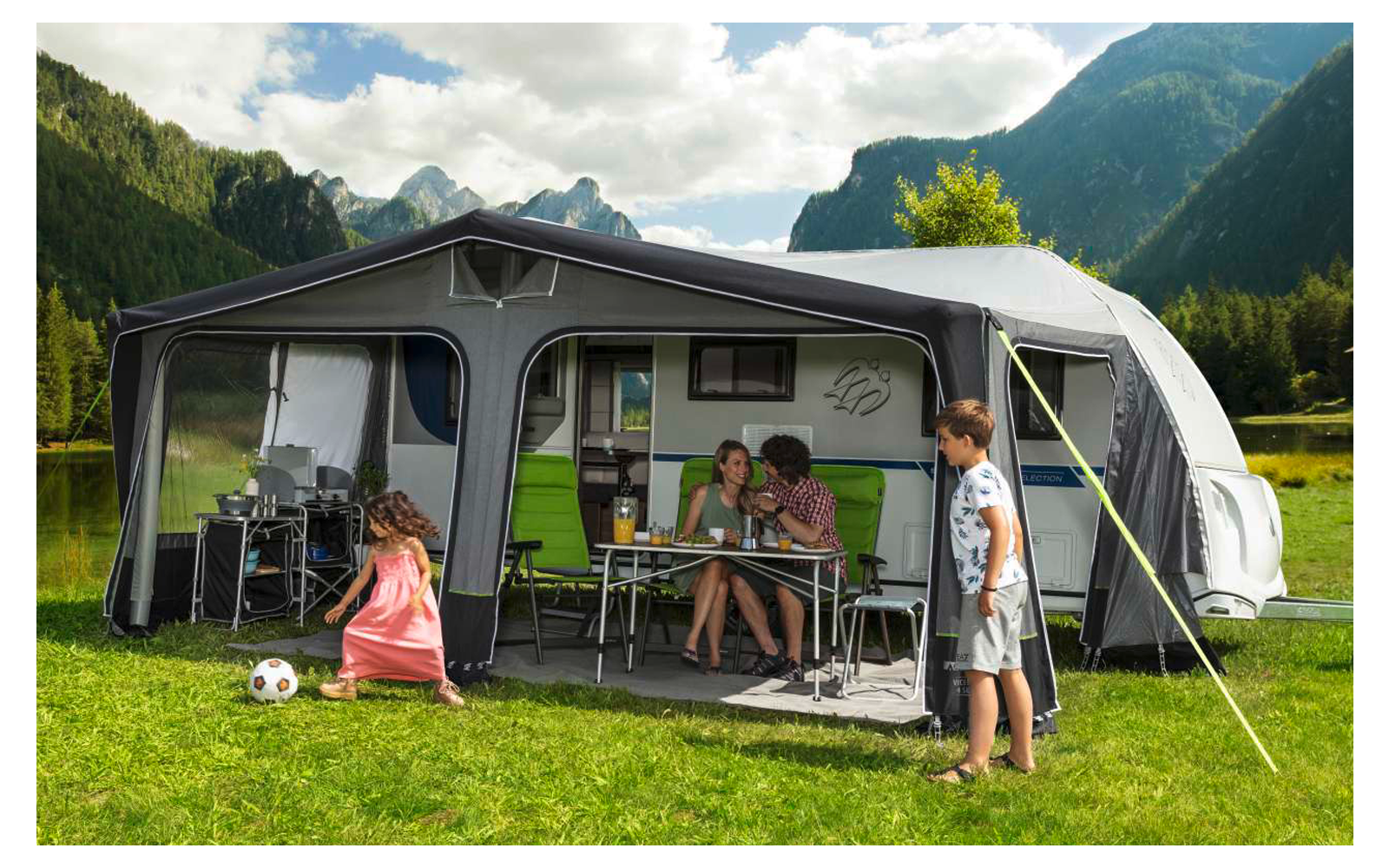 People sitting and standing in front of a caravan with an attached awning on a green meadow in the mountains